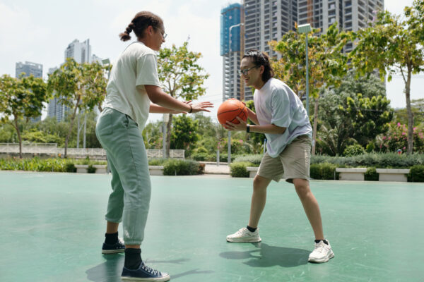 Two people enjoying basketball game on spacious outdoor court surrounded by greenery and modern buildings in background