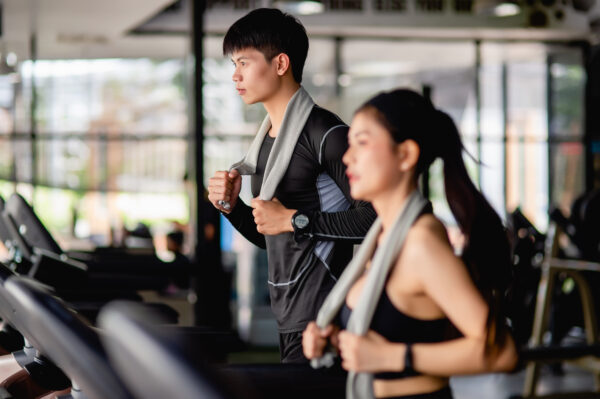 Selective focus young man, blurred young sexy woman in foreground wearing sportwear and smartwatch, They are running on treadmill to for workout in modern gym, copy space